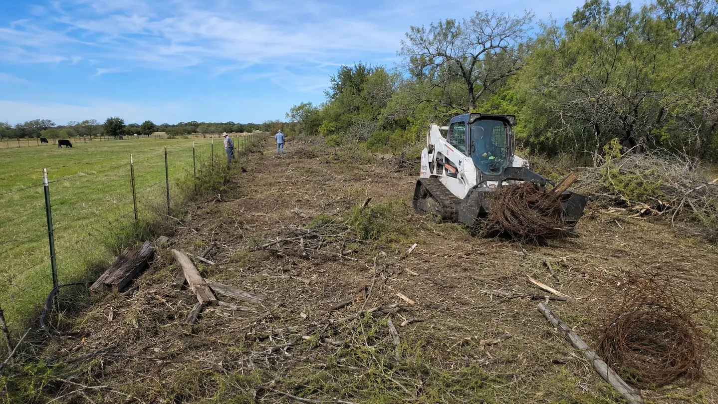 Compact equipment clearing brush, old wire, and broken posts along a rural fence line.